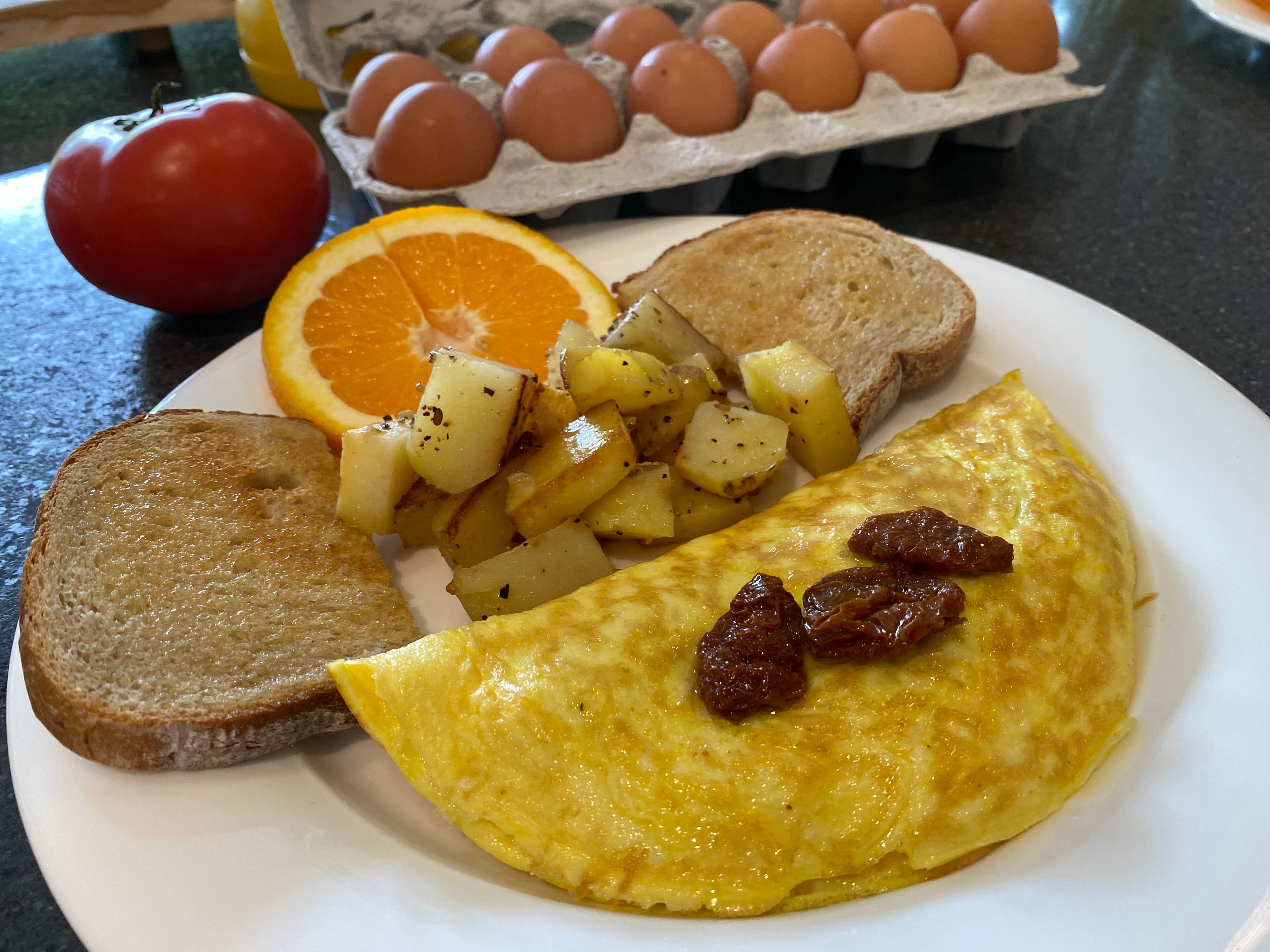 A plate featuring an omelet topped with sun-dried tomatoes, alongside toast, fried potatoes, an orange slice, and a tomato, with a carton of eggs in the background.